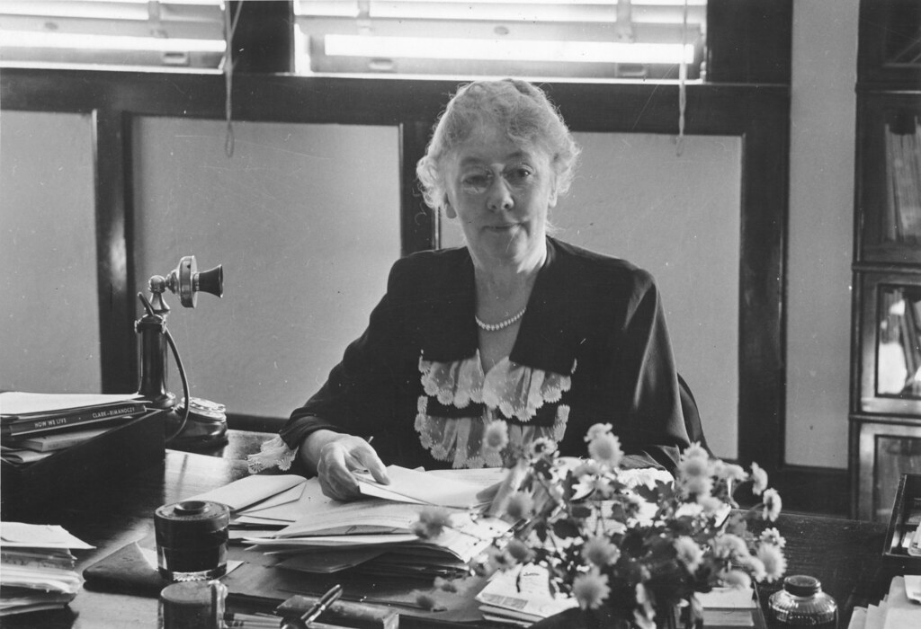 A black and white photo of Mary Alice Baldwin sitting behind a cluttered desk with an old phone and flowers on it