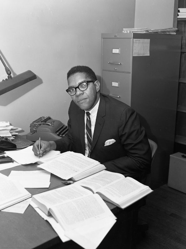 Cook looks up from a desk in a small office. The desk is covered with papers and books, and he holds a pen over a document his is writing on.