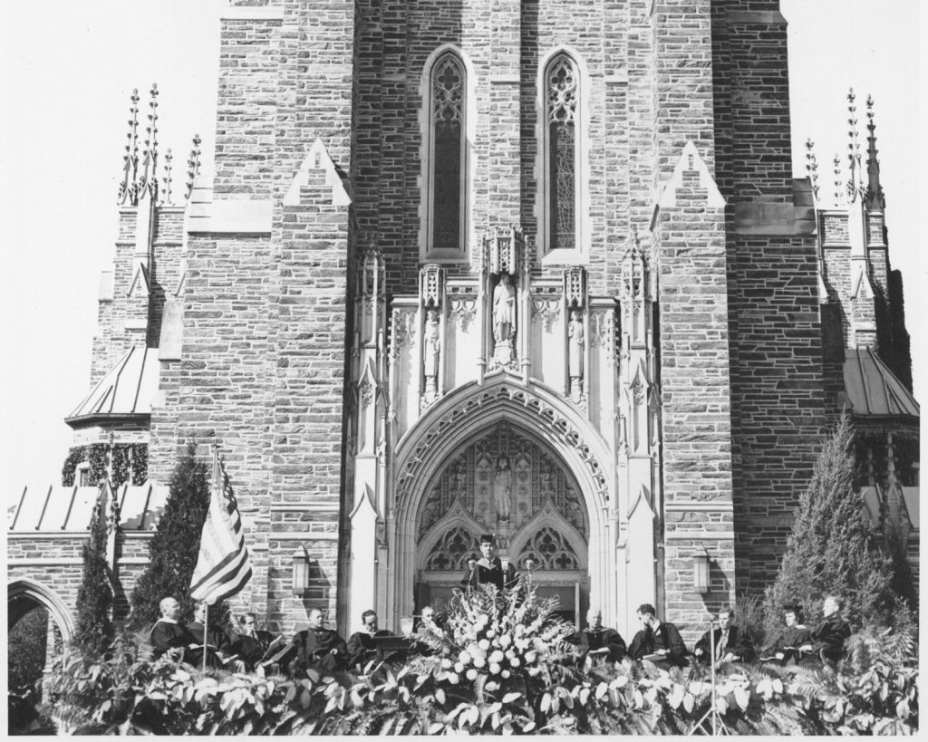 Edens standing at a podium with flowers in front of it in front of Duke Chapel 