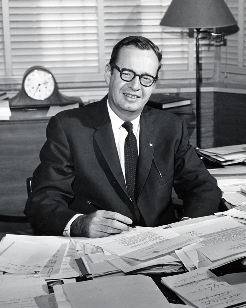 President Douglas Knight sitting at a desk covered in papers