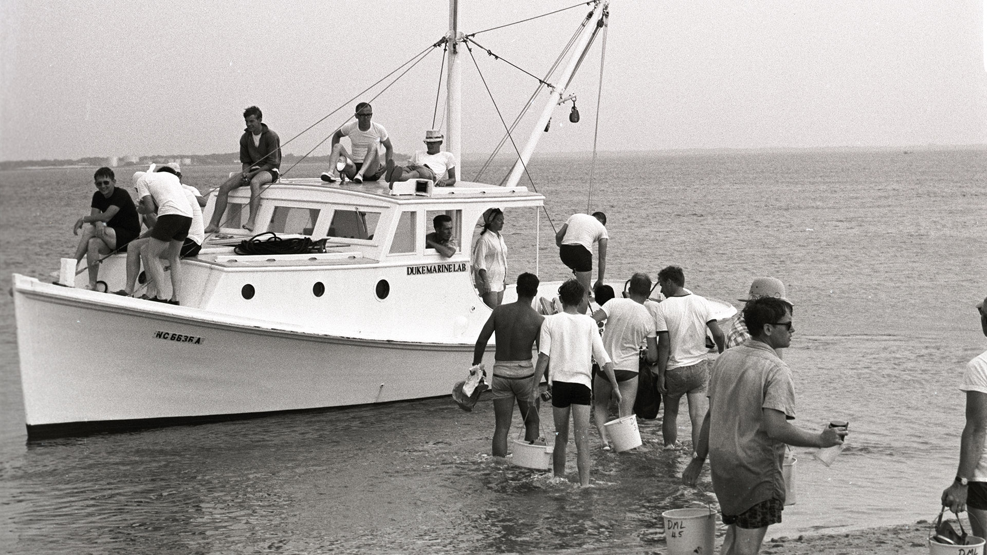 A black and white photo of people walking out to board a white boat