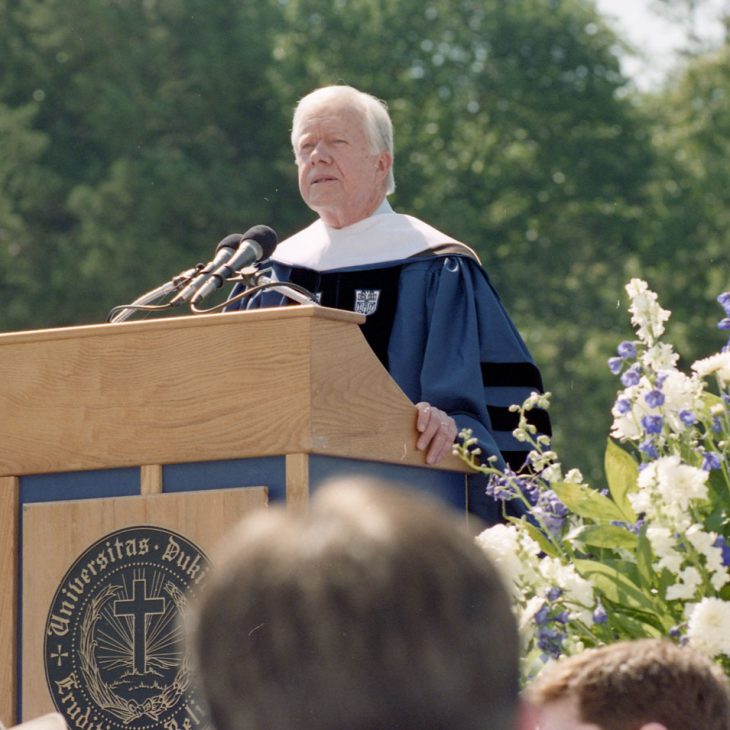 President Jimmy Carter speaking at the podium at commencement with flowers in the foreground and the stands of Wallace Wade Stadium in the background
