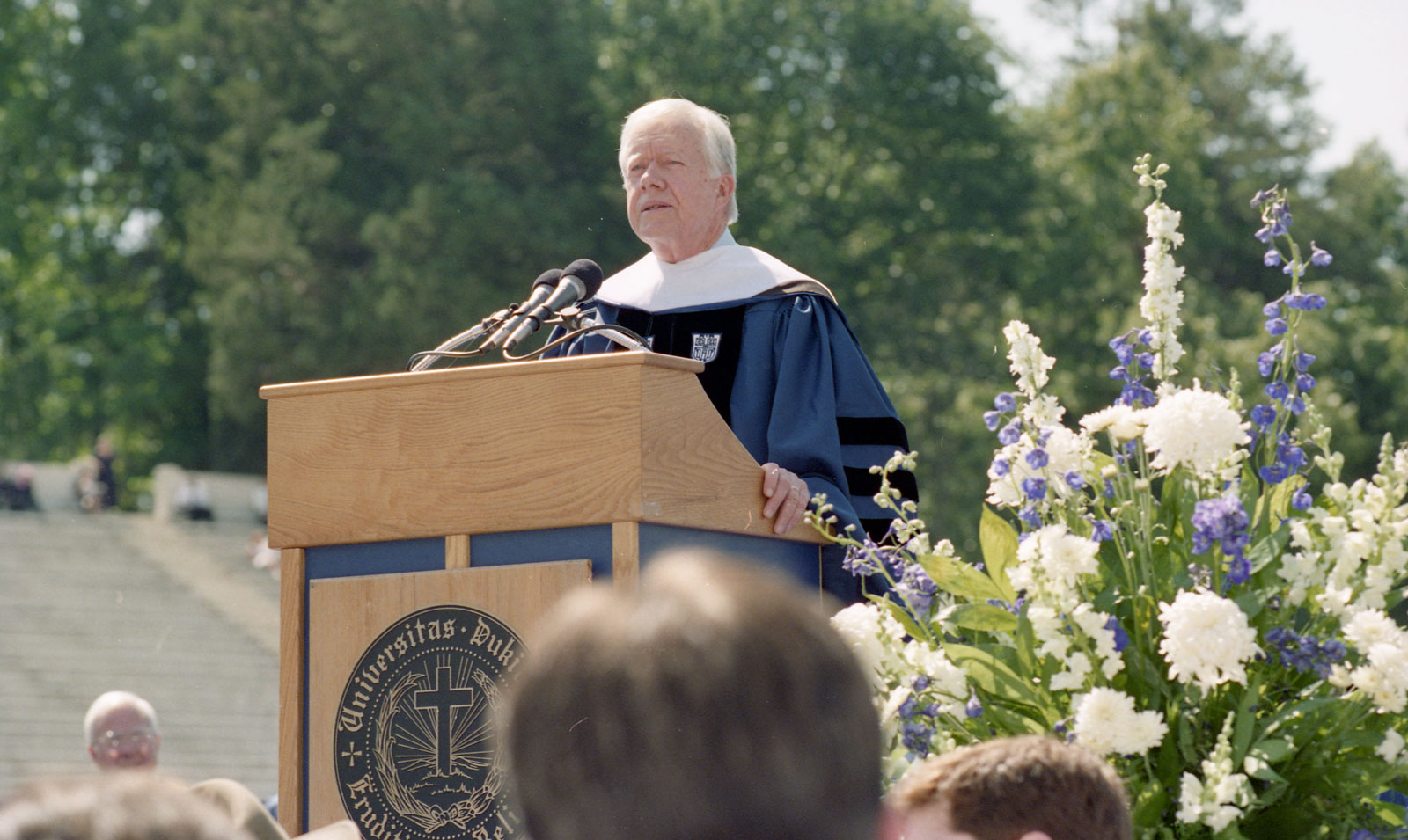 President Jimmy Carter speaking at the podium at commencement with flowers in the foreground and the stands of Wallace Wade Stadium in the background