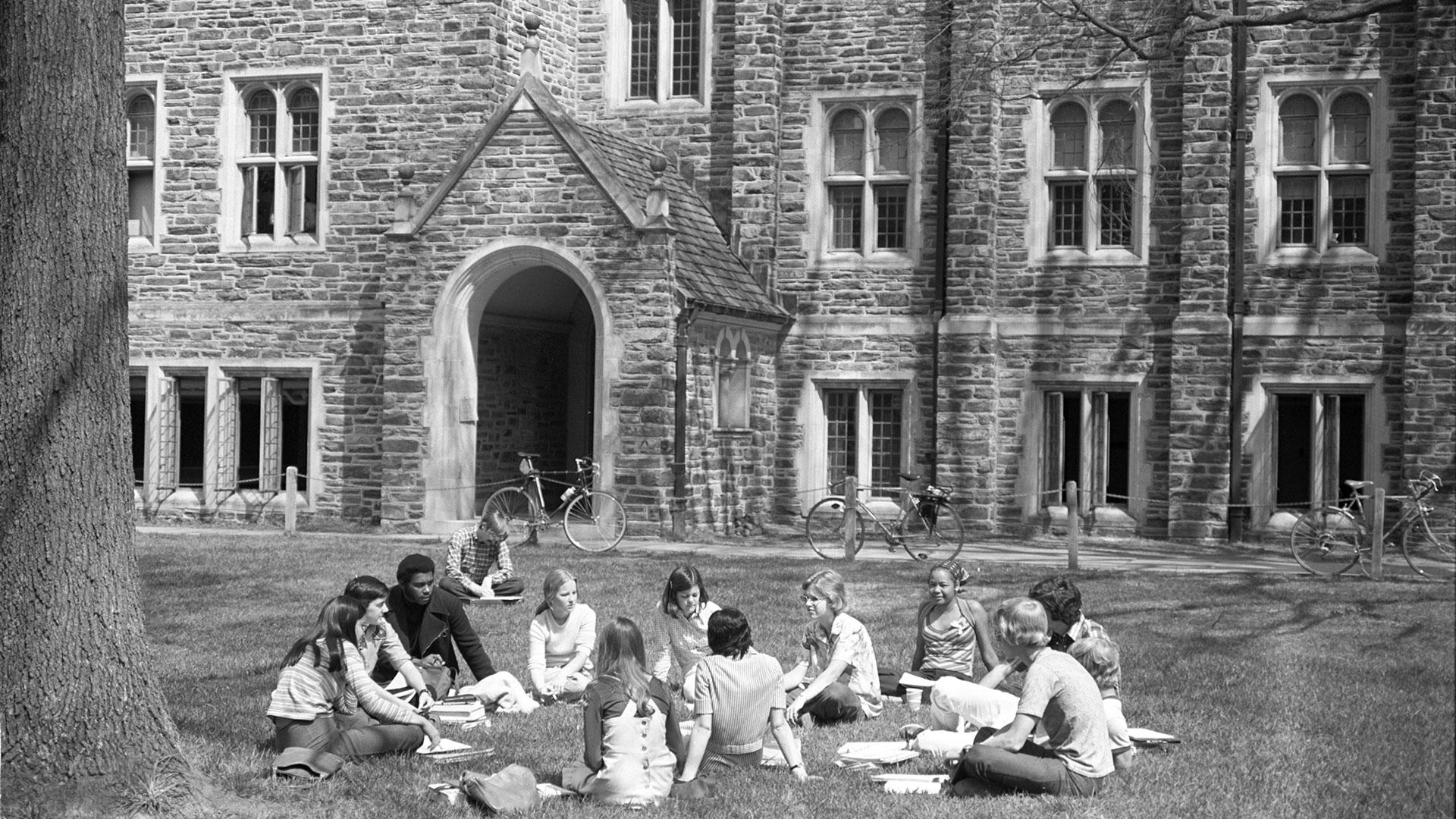 Students sit in a circle on the grass next to a tree in front of the Old Chemistry building