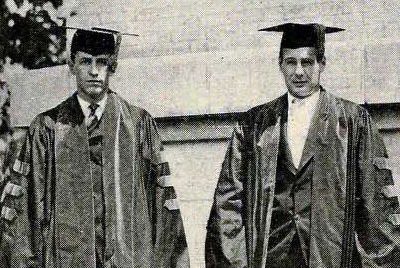 Two students in doctoral robes stand in front of a brick wall