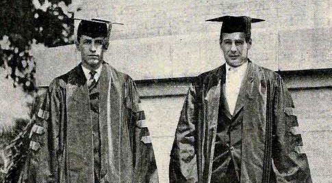 Two students in doctoral robes stand in front of a brick wall