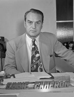 Eddie Cameron in a suit and tie sitting at a desk