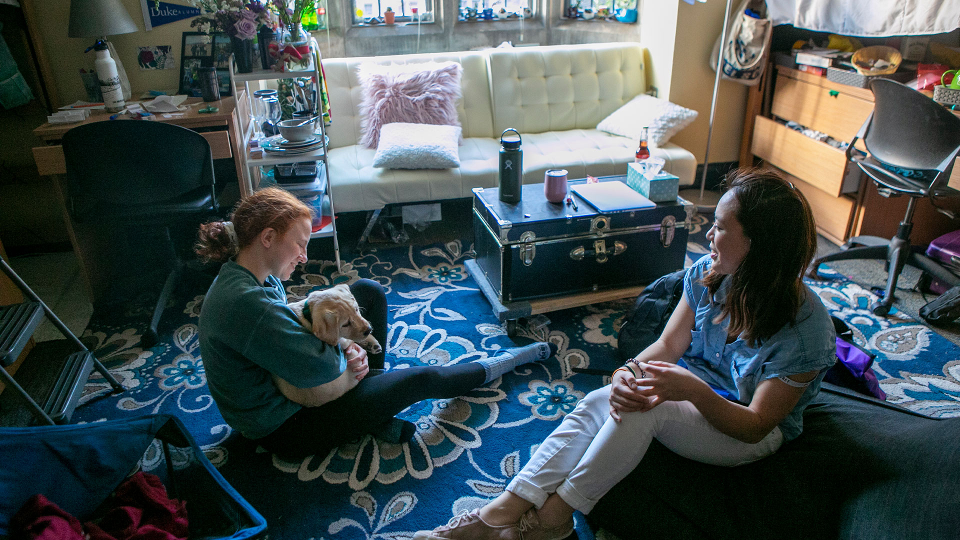 Two women sit in a dorm room with a carpet, futon, ottoman fully decorated while holding a puppy