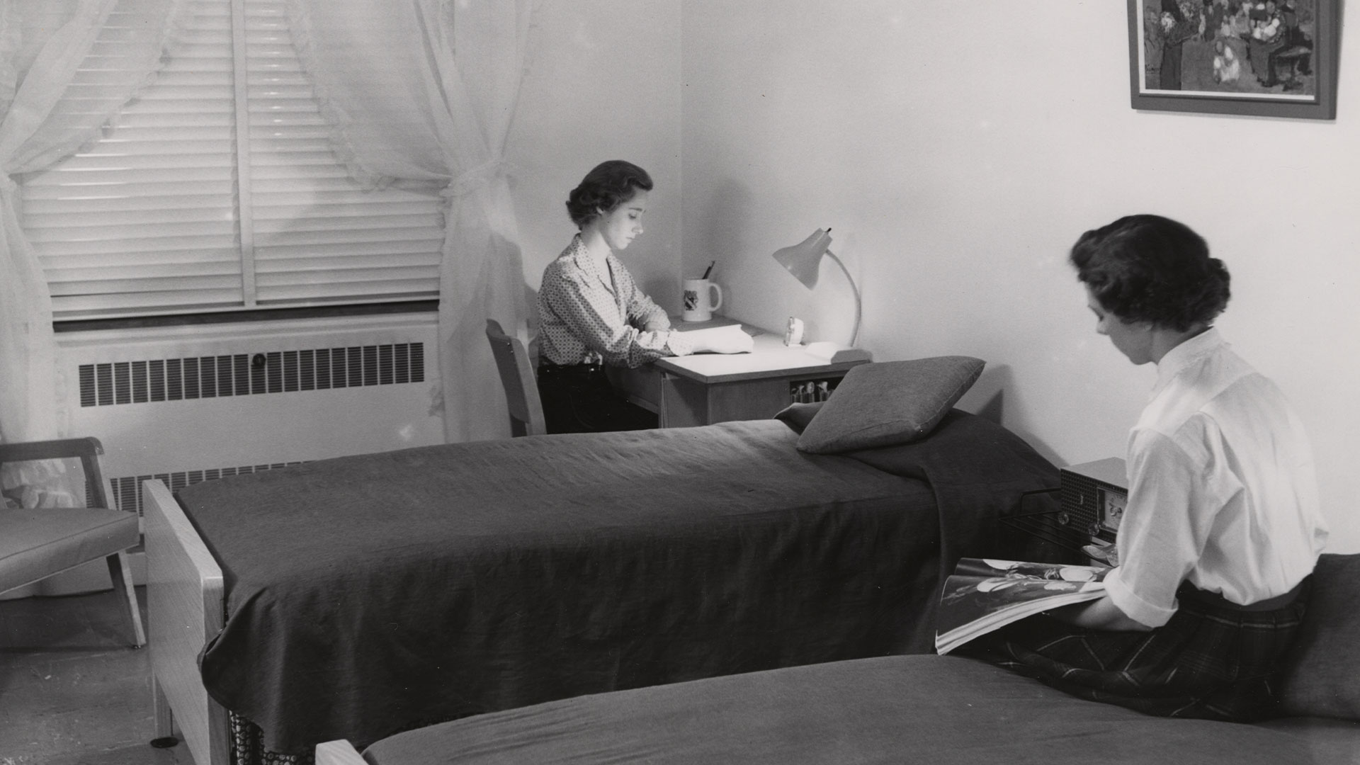 Two women sit in a dorm room with 2 beds and a desk