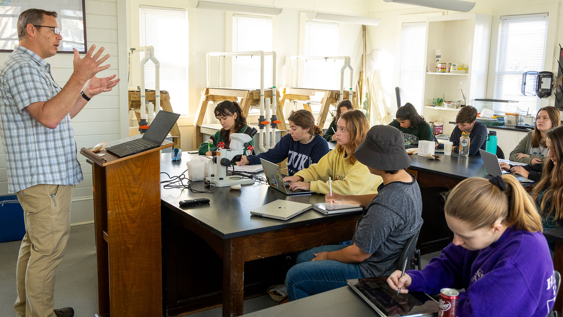 A current photo of students in a classroom listening to an instructor at the podium in a well-lit room