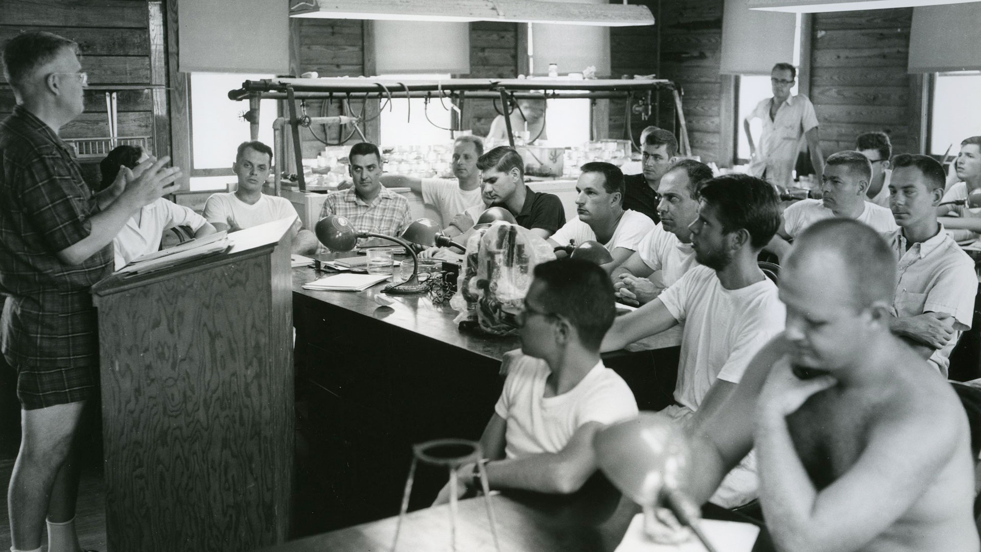 An old photo of male students in a dark classroom listen to an instructor at a podium