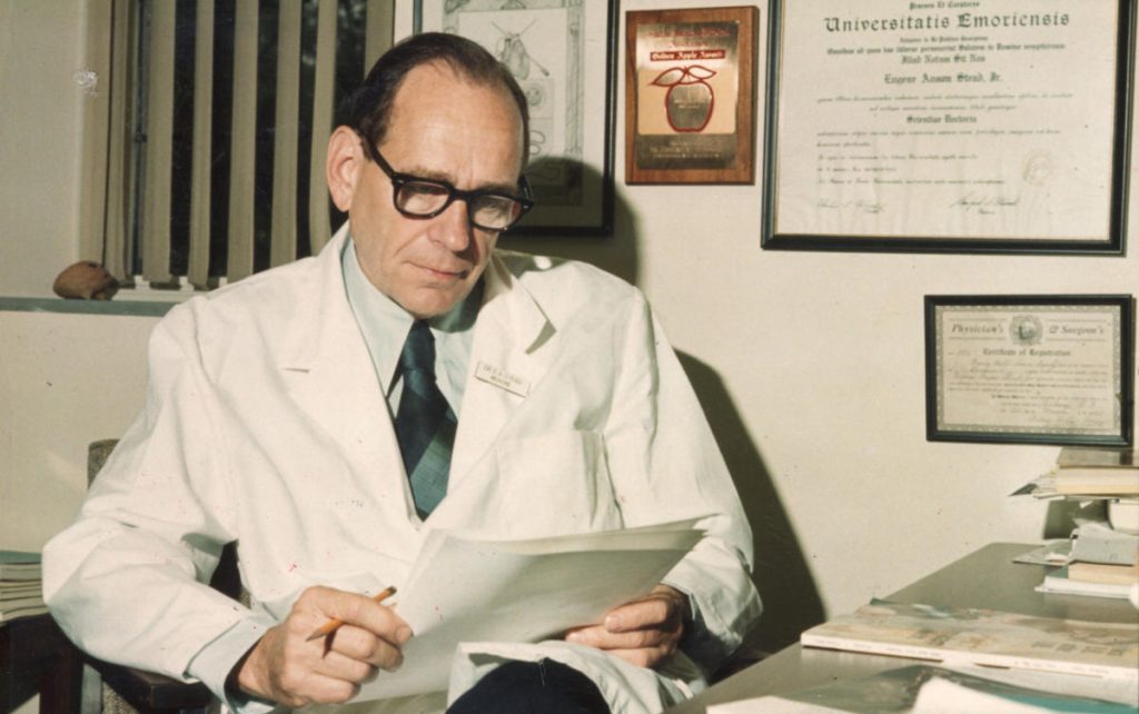 An old photo of Eugene Stead sitting at a desk in his office wearing a lab coat looking over paperwork