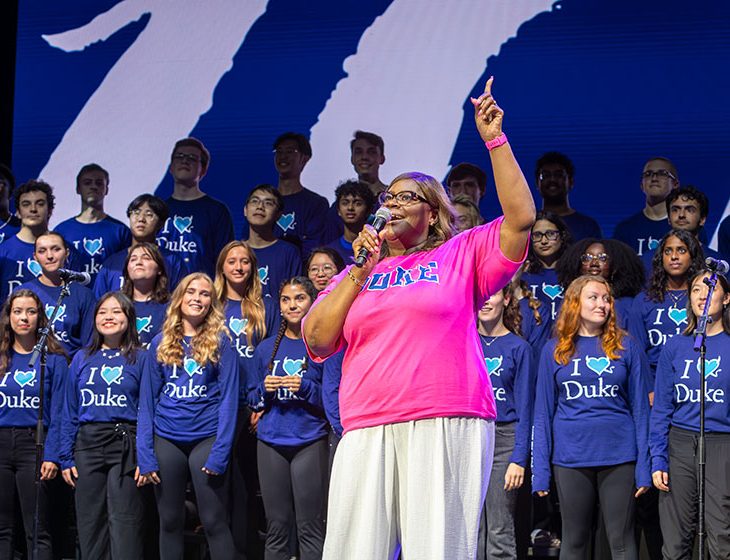 Retta holds up 1 finger pinting "higher" as the Duke Chorale gets ready to sing in the background